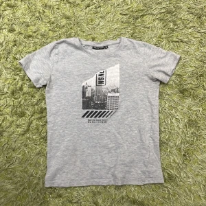 Grå t-shirt med tryck från Reaction - Säljer en grå t-shirt från Reaction med ett coolt tryck av en stadssilhuett och texten 'Next Stop Urban District New York'. Perfekt för en urban look. T-shirten är kortärmad och gjord i mjukt material.