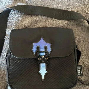 Trapstar väska  - Snygg svart axelväska från Trapstar. Väskan har en justerbar axelrem med märkeslogga. Perfekt för att bära dina nödvändigheter, finns mycket utrymme. Egna bilder. Fick i present gissar att priset ligger på över 1200kr men säljer den för mycket billigare!