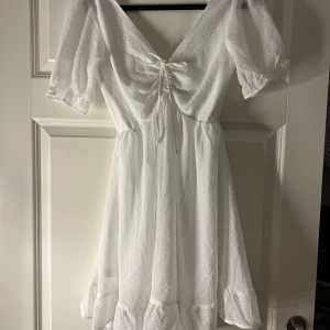 Vit studentklänning med volangdetaljer - Helt ny vit studentklänning med prislapp med volangdetaljer och knytning framtill och bak. Klänningen har korta ärmar och en v-ringad design. Klänningen är i storlek 32 men är stor i storlek så passar mig som vanligtvis har 36/S. 