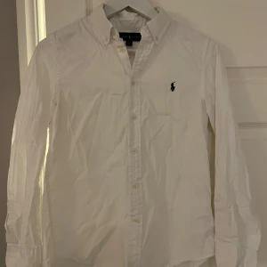 Vit skjorta från Ralph Lauren - Snygg vit skjorta från Ralph Lauren med klassisk krage och broderad logga på bröstet. Skjortan har långa ärmar och knappar framtill. Perfekt för en stilren look. Storlek S