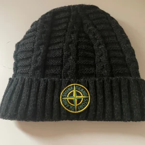 Grå stickad mössa från Stone Island - Snygg grå stickad mössa från Stone Island med ribbad kant och ett broderat märke framtill. Perfekt för kyliga dagar och ger en stilren look.