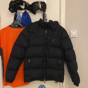 dunjacka från Polo Ralph Lauren - Säljer en svart dunjacka från Polo Ralph Lauren med dragkedja och huva. Jackan har en broderad logotyp på bröstet och praktiska fickor med dragkedja. Perfekt för kyliga dagar knappt använd.