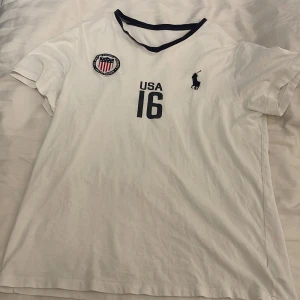 Vit t-shirt  - Skön vit t shirt med en broderad hästpolo ryttare och en usa flagga. Står USA och nummer 16 på framsidan. 100% bomull. Vet inte storlek men gissar på M!!