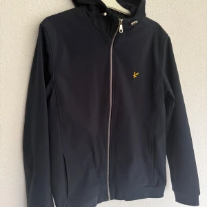 Svart jacka från Lyle & Scott - Säljer en stilren marin blå jacka från Lyle & Scott med dragkedja och huva. Jackan har en broderad logotyp på bröstet och är perfekt för kyligare dagar. Den är långärmad och har en modern passform.