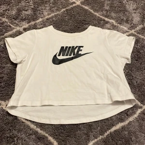 Vit croppad t-shirt från Nike - Säljer en vit croppad t-shirt från Nike med det klassiska logotyptrycket i svart på framsidan. Perfekt för en sportig och avslappnad stil. T-shirten har korta ärmar och rund halsringning.