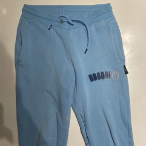 Ljusblå mjukisbyxor från Hoodrich - Säljer ett par ljusblå mjukisbyxor från Hoodrich i storlek XS. Byxorna har en bekväm passform med snörning i midjan och en broderad logga på benet. Perfekta för en avslappnad stil. Paras också ihop med hoodien 