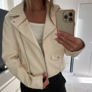 Vit skinnjacka  - Säljer en snygg vit skinnjacka från BERSHKA som är perfekt för höst och vår. Denna jackan är i utmärkt skick och är i storlek M men passar bättre som en XS/S. Jackan har en praktisk dragkedja framtill och två silverknappar som tillför en extra detalj
