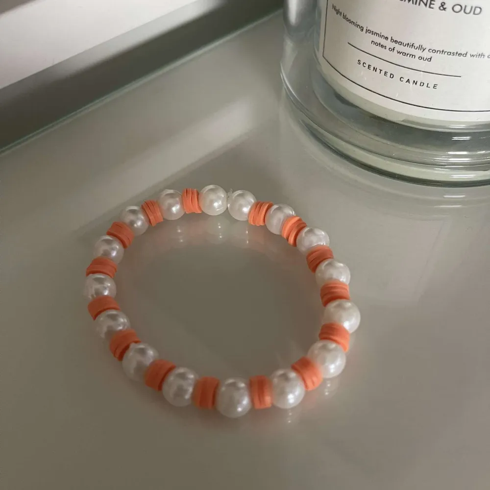 Snyggt armband med vita pärlor och orangea detaljer. Perfekt för att ge en färgklick till din outfit. Armbandet är elastiskt och passar de flesta handleder.. Accessoarer.