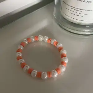 Snyggt armband med vita pärlor och orangea detaljer. Perfekt för att ge en färgklick till din outfit. Armbandet är elastiskt och passar de flesta handleder.