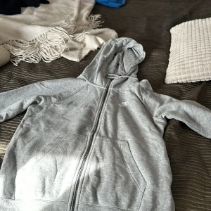 Grå fleecejacka från Nike - Säljer en supermjuk och bekväm grå fleecejacka från Nike. Den har en dragkedja framtill och en praktisk huva. Perfekt för kyliga dagar eller som ett extra lager. Jackan har långa ärmar och två fickor framtill. Passar både till vardags och träning!