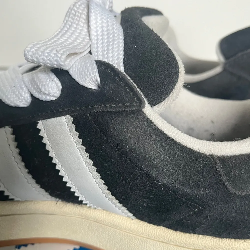 Säljer ett par svarta Adidas Campus sneakers i mocka med vita detaljer. Skorna har de klassiska tre ränderna på sidan och en vit sula. De är i bra skick med lite slitage på sulan. Perfekta för vardagsbruk och ger en stilren look.. Skor.