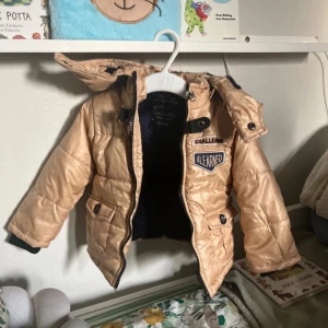 Beige pufferjacka för barn från Lay Lay - Säljer en beige pufferjacka för barn från Lay Lay. Jackan har en mysig mörkblå insida och en avtagbar huva. Den är försedd med dragkedja och knappar framtill samt två fickor med knappar. Perfekt för kyligare dagar!