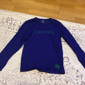 Blå långärmad tröja från Calvin Klein - Säljer en stilren blå långärmad tröja från Calvin Klein med grönt tryck på bröstet. Tröjan har en rund halsringning och en liten grön etikett med CK-logga nedtill. Perfekt för en avslappnad look. Kan gå ner i pris vid snabb affär🫶