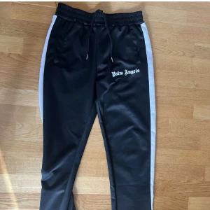 Svarta träningsbyxor från Palm Angels - Snygga svarta träningsbyxor från Palm Angels med vita sidoremsor och logga på låret. Byxorna har en elastisk midja med snörning för bästa passform. Perfekta för en sportig look eller avslappnad stil.