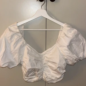 Vit kortärmad topp från Gina Tricot - Säljer en söt vit topp från Gina Tricot med korta puffärmar och rynkdetaljer framtill. Perfekt för en somrig look. Toppen har en elastisk kant för en bekväm passform.