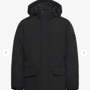 Svart parkajacka från Tommy Hilfiger - Snygg svart parkajacka från Tommy Hilfiger med huva och diskret logotyp på ärmen. Jackan har långa ärmar och praktiska fickor framtill. Perfekt för kyliga dagar. Fickor uppe vid jackan samt utanför fickorna och även fickorna som syns på bilden. Sparsamt använd nypris 4600 kr 