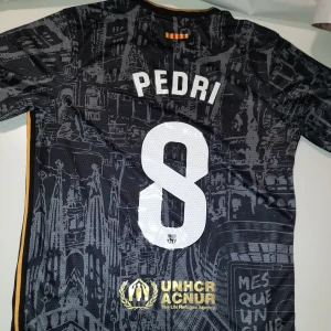 Pedri Barcelona Fc Tröja  - Snygg svart fotbollströja från Nike med guldiga detaljer och tryck. Tröjan har korta ärmar och ett unikt mönster över hela tyget. Den har sponsorloggor och spelarens namn och nummer på ryggen. Perfekt för fotbollsfans! PEDRI/8