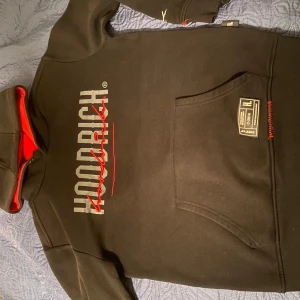 Svart hoodie från Hoodrich - Storlek S, Säljer en svart hoodie från Hoodrich med röd insida och tryck på framsidan. Hoodien har en praktisk magficka och detaljer i rött. Perfekt för streetwear-stilen.
