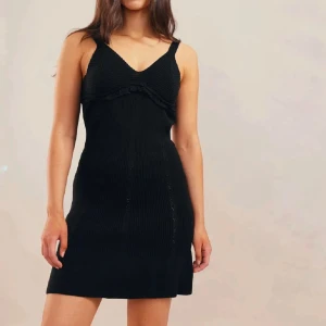 Svart stickad klänning - Snygg svart stickad klänning med breda axelband och volangdetalj vid bysten. Klänningen har en figurnära passform och en subtil ribbad struktur som ger en elegant look. Perfekt för en stilren outfit.