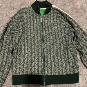 Lacoste monogram - Säljer en grön mönstrad monogram tröja från Lacoste med dragkedja och ribbad krage. tröjan har ett geometriskt mönster och en vit rand längs ärmarna. Perfekt för en sportig look!