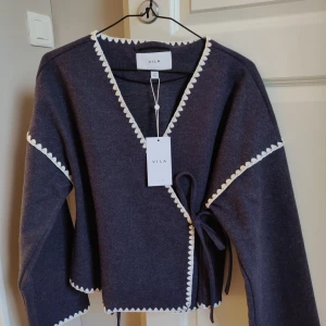 Mörkgrå kofta från Vila - Säljer en elegant mörgrå kofta från Vila med vita spetsdetaljer längs kanterna. Koftan har en knytning i sidan och långa ärmar, perfekt för en stilren look. Passar utmärkt till både vardag och speciella tillfällen.