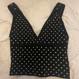 Svart prickig topp - Säljer en svart prickig topp med v-ringning. Toppen har breda axelband och ett stilrent mönster med vita prickar. Perfekt för en trendig look!