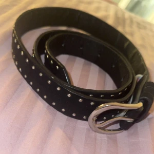 Svart bälte med nitar - Snyggt svart bälte dekorerat med små silverfärgade nitar. Perfekt för att ge en edgy touch till din outfit. Bältet har ett klassiskt spänne i silver och är justerbart för bästa passform.