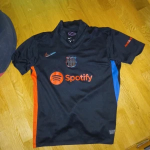 Svart FC Barcelona fotbollströja - Snygg svart FC Barcelona tröja med korta ärmar och sponsorloggor från Spotify och UNHCR. Tröjan har spelarnamnet R. Araujo och nummer 4 på ryggen. Tillverkad i Dri-FIT material för optimal komfort.