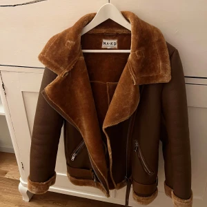 Brun pälsjacka från NA-KD storlek 38 - Säljer en snygg brun pälsjacka från NA-KD med mjukt foder och bred krage. Jackan har dragkedjor och en stilren design som passar perfekt för kyliga dagar. Perfekt för dig som vill ha både stil och värme! Storlek 38.