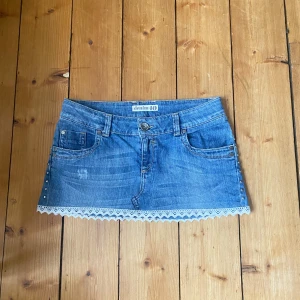 Blå jeanskjol med spetsdetaljer - Snygg blå jeanskjol från Denim med spetskant och nitar längs sidorna. Kjolen har klassiska femficksdetaljer och low waist. Perfekt för en cool outfit med en touch av elegans. Omsydd från jeans, fint gjort! 