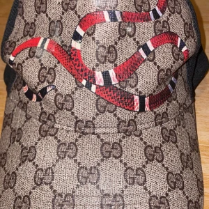 Mönstrad keps med orm - Snygg keps med ett coolt ormmönster i rött och vitt på en brun bas med logotypmönster. Baksidan är i svart meshmaterial för bra ventilation. Perfekt för att ge din outfit en unik touch.