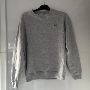 Grå sweatshirt från Lacoste - Säljer en klassisk grå sweatshirt från Lacoste med rund hals och det ikoniska krokodilmärket på bröstet. Tröjan har långa ärmar och ribbade muddar vid ärmslut och nederkant. Perfekt för en avslappnad stil.