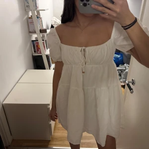 Vit klänning från Zara - Charmig vit klänning från Zara med puffiga kortärmar och en söt knytning framtill. Perfekt för varma dagar med sin luftiga design och korta längd. Klänningen har en romantisk känsla och är lätt att styla för olika tillfällen.