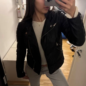 Svart skinnjacka med dragkedjor - Snygg svart skinnjacka med coola dragkedjedetaljer och bälte i midjan. Jackan har en klassisk bikerstil med quiltade axlar och långa ärmar. Perfekt för en edgy look!