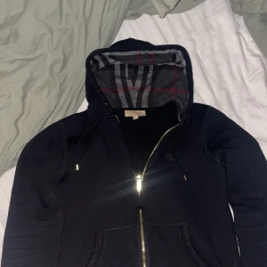 Svart hoodie från Burberry - Snygg svart hoodie från Burberry med dragkedja och broderad logga på bröstet. Insidan av huvan har ett klassiskt rutigt mönster i grått och rött. Perfekt för en stilren look.