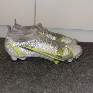Nike Mercurial fotbollsskor i silver och gult - Snygga Nike Mercurial fotbollsskor med glittrig silverdesign och gula detaljer. Skorna har en strömlinjeformad passform och snörning för optimal kontroll på planen. Perfekta för den som vill sticka ut med stil.