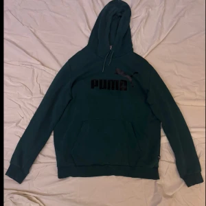 Grön hoodie från Puma - Säljer en snygg grön hoodie från Puma med deras ikoniska logga i svart på bröstet. Tröjan har en klassisk känguruficka och justerbar huva med dragsko. Perfekt för en avslappnad stil.