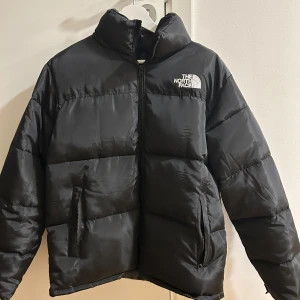 Svart dunjacka från The North Face - Säljer en svart dunjacka från The North Face med dragkedja och broderad logga på bröstet. Jackan har långa ärmar och är perfekt för kalla dagar. Den är designad för att hålla dig varm och bekväm med sin pufferstil.