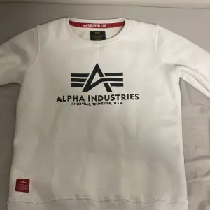Säljer en stilren vit sweatshirt från Alpha Industries med deras ikoniska logga tryckt på framsidan. Tröjan har långa ärmar och en rund halsringning. Perfekt för en avslappnad look.