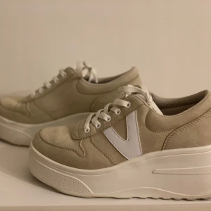 Beige sneakers med vit detalj - Snygga beige sneakers med vit sula och vit V-detalj på sidan. Skorna har snörning och en bekväm, upphöjd sula för extra komfort. 