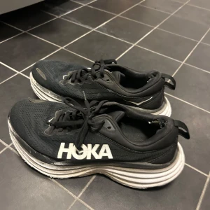 Svarta löparskor från Hoka - Säljer ett par svarta löparskor från Hoka med vit sula och snörning. Skorna har en sportig design och är perfekta för löpning och träning. Lite slitna i hälen 