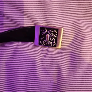 Svart bälte från Tiger of Sweden - Snyggt svart bälte från Tiger of Sweden med en unik spänne-design. Perfekt för att ge en stilren touch till din outfit. Bältet är tillverkat i skinn och har en elegant finish.