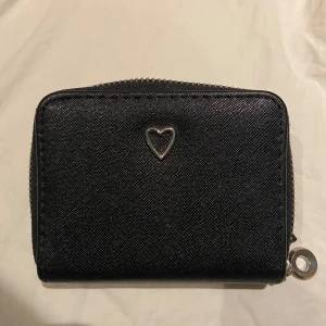 Svart plånbok med hjärtdetalj - Snygg svart plånbok med en söt hjärtdetalj på framsidan. Plånboken har en praktisk dragkedja runtom och flera fack för kort och mynt. Perfekt för att hålla ordning på dina tillhörigheter.