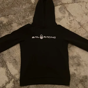Sail Racing - Snygg svart hoodie från Sail Racing med vit logga på bröstet. Perfekt för en avslappnad stil med en klassisk look. Den har en bekväm huva och långa ärmar. Tröjan är i bra skick.  Obs! Det är barnstorlek 