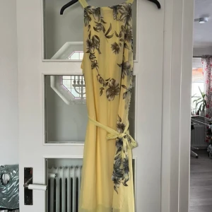 Klänning - Elegant gul långklänning med vackert blommönster i grå/brun toner. Klänningen har en knytning i midjan, vilket ger en luftig och somrig känsla. Perfekt för speciella tillfällen. Storlek S men passar även M. Klänningen har fina blommor. Den passar perfekt för sommaren om man vill vara lite finklädd! 🌾🌼🌷 Klänningen är i nytt skick och har bara använts 1 gång! Kontakta mig gärna för mer information eller bilder!🍄‍🟫❤️