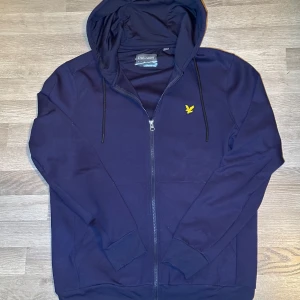 Mörkblå zip hoodie från Lyle & Scott - Säljer en stilren mörkblå hoodie från Lyle & Scott med dragkedja och huva. Den har en gul logotyp på bröstet och justerbara snören vid huvan. Perfekt för alla årstider. Priset är inte hugget i sten. Kontakta mig vid övriga frågor 