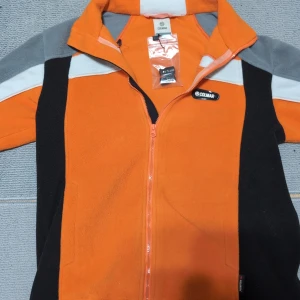 Orange fleecejacka från Colmar - Tja Säljer en l orange fleecejacka från Colmar med svarta och grå detaljer. Jackan har en dragkedja framtill och är perfekt för kyliga dagar. Den har en sportig look med långa ärmar och en bekväm passform. Priset kan diskuteras vid en snabb affär dm för mer frågor och bilder