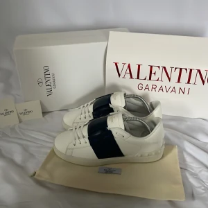 Valentino Garavani sneakers i vitt och blått - Snygga Valentino Garavani sneakers i vitt med en bred blå rem över ovansidan. Skorna har en stilren design med snörning och är tillverkade i skinn. Perfekta för en trendig look.