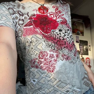 Mönstrad t-shirt med röda detaljer - Säljer en unik t-shirt med ett mixat mönster i grått och beige, prydd med röda och svarta detaljer. T-shirten har korta ärmar och en avslappnad passform. Perfekt för dig som vill sticka ut med en kreativ stil! Storlek XL men jag använder som översized T-shirt. Skulle även vara bra för att sy om då det är ett väldigt fint mönster.