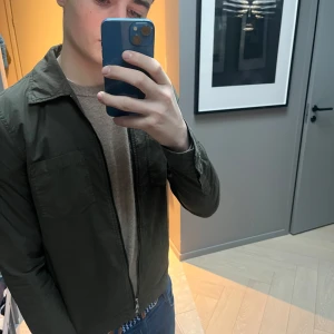 Olivgrön jacka från Oscar Jacobson, windbraker  - Snygg olivgrön jacka från Oscar Jacobson med dragkedja och krage. Jackan har två bröstfickor och långa ärmar med knappdetaljer. Perfekt för en stilren look. Nypris 2500kr